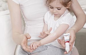 Bambina piccola seduta sul grembo della mamma, che sta applicando Eucerin AtopiControl Crema Fasi Acute sulla piega del braccio.